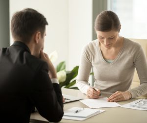 Satisfied with loan terms woman singing contract when sitting at desk in front of male bank employee at office. Millennial female applicant accepting employers offer and putting signature in agreement